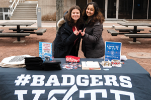 Two girls stand behind a black table with #BadgersVote printed on the front, holding up the Wisconsin W with their hands.